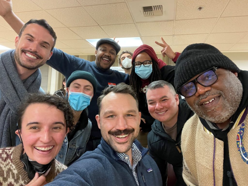 A group of nine people of different ages, races and genders are grinning into the camera. 