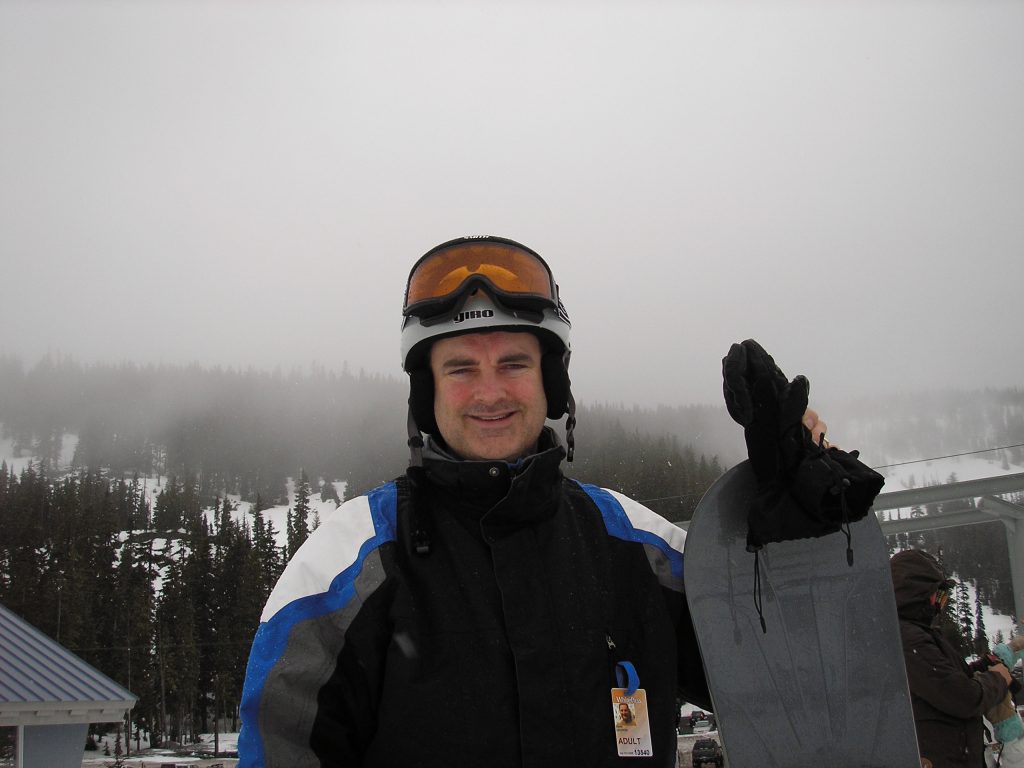A photo of Mark Gropper smiling with a snowboard and snowy mountains in the background. 